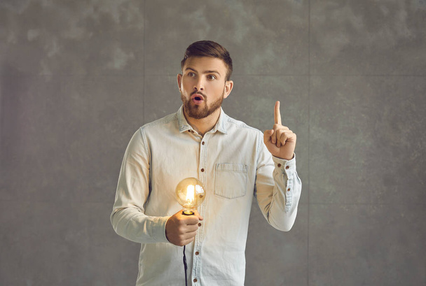 Surprised man thinking and holding glowing lightbulb has bright idea studio shot - Photo, image