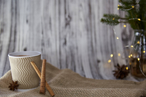 Wooden and sackcloth organic Christmas background design with copy space. Cardboard cup with hot beverage, cinnamon sticks and anise star. Christmas tree with garlands in the background - Photo, Image