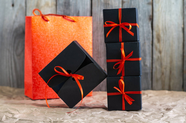 Gifts in black boxes tied with a red ribbon and a red shopping bag stand on craft paper against a wooden background - Photo, image