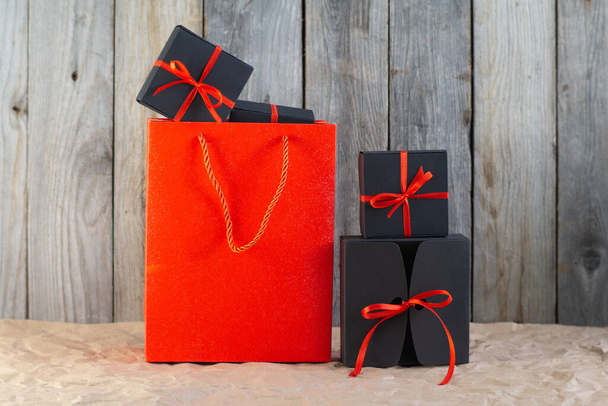 Gifts in black boxes tied with a red ribbon and a red shopping bag stand on craft paper against a wooden background - Фото, изображение
