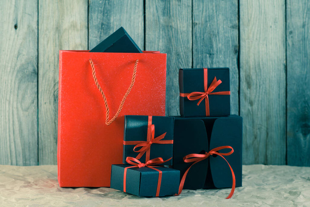 Gifts in black boxes tied with a red ribbon and a red shopping bag stand on craft paper against a wooden background - Foto, afbeelding