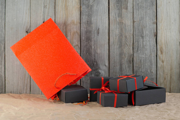 Gifts in black boxes tied with a red ribbon and a red shopping bag stand on craft paper against a wooden background - Foto, imagen