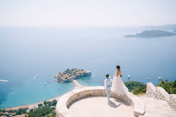 The bride stands on the parapet of the observation deck overlooking the island of Sveti Stefan, the groom holds her hand - Fotoğraf, Görsel