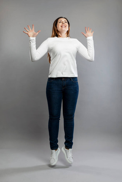 Adult woman jumping. Smiling brunette in jeans and a white sweater. Happiness and positivity. Gray background. Vertical. - Фото, зображення