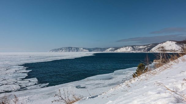 Il fiume Angara, privo di ghiaccio, fuoriesce dal lago ghiacciato Baikal. Il ghiaccio sciolto galleggia sull'acqua blu. Minuscole sagome di persone sul ghiaccio. Neve ed erba secca sulla riva. Montagne contro il cielo azzurro. - Foto, immagini