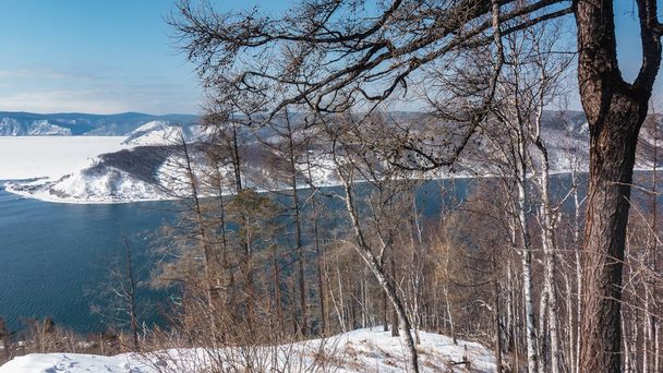 O rio Angara sem gelo azul flui entre as margens cobertas de neve. Árvores nuas contra o céu azul e montanhas. Uma seção de congelado Lago Baikal é visível. Sibéria. Listvyanka - Foto, Imagem
