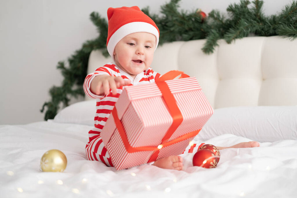 Niña en traje de cuerpo a rayas y Santa sombrero jugando con caja de regalo en el dormitorio decorado de Navidad - Foto, Imagen