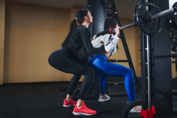 Genç esmer kadın spor salonunda halterle bacak çalışıyor. Kişisel kadın eğitmen. Çömelme egzersizi  - Fotoğraf, Görsel