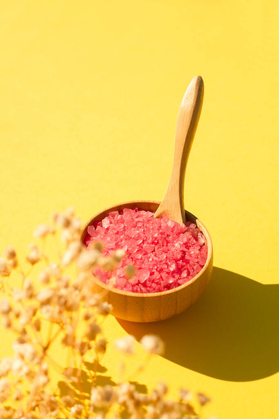 Wooden bowl with a spoon filled with pink bath sea salt. Beauty treatment for spa and wellness on yellow background. Skincare natural cosmetic concept for body care with dry plants and shadows - Foto, Imagem