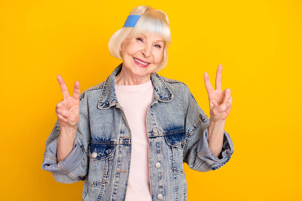 Photo portrait of senior woman smiling cheerful in denim outfit showing v-sign gesture isolated on bright yellow color background - Photo, image