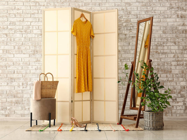 Beige folding screen with yellow dress, mirror and pouf near light brick wall - Photo, Image
