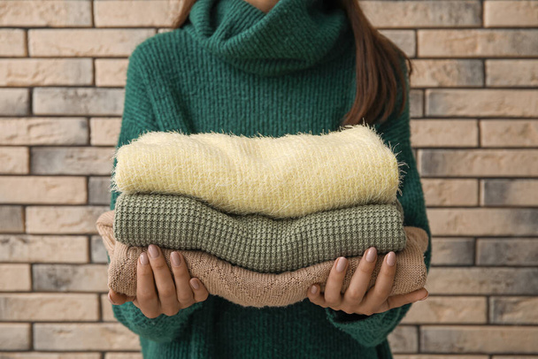 Mujer joven sosteniendo pila de suéteres modernos suaves sobre fondo de pared de ladrillo - Foto, Imagen