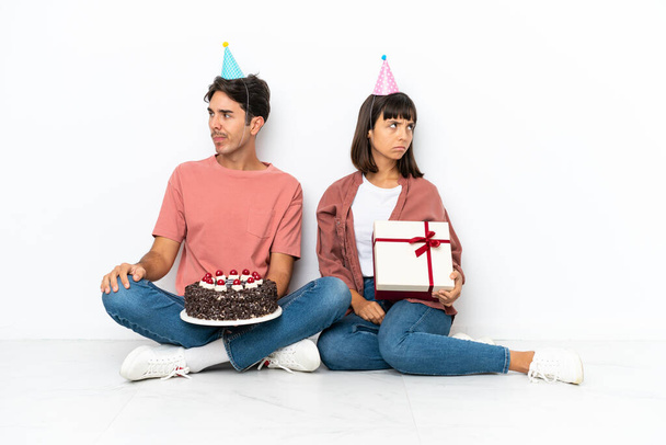 Jovem casal misto comemorando um aniversário sentado no chão isolado no fundo branco com expressão facial confusa enquanto morde lábio - Foto, Imagem