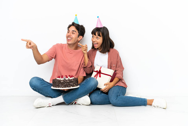 Pareja joven de raza mixta celebrando un cumpleaños sentada en el suelo aislada sobre fondo blanco apuntando hacia un lado para presentar un producto - Foto, imagen