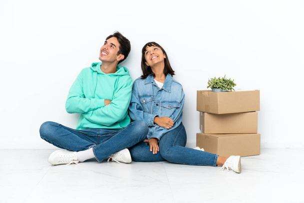 Молодая пара делает шаг, поднимая коробку, полную вещей, сидя на полу изолированы на белом фоне глядя вверх, улыбаясь - Фото, изображение