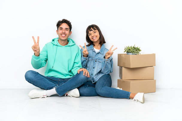 Молодая пара делает шаг, поднимая коробку с вещами, сидя на полу изолированы на белом фоне улыбаясь и показывая знак победы обеими руками - Фото, изображение