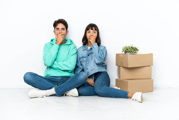 Молодая пара делает шаг, поднимая коробку с вещами, сидя на полу изолированы на белом фоне покрывая рот руками за то, что сказал что-то неуместное - Фото, изображение