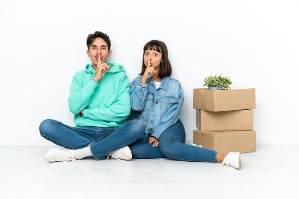 Молодая пара делает шаг, поднимая коробку с вещами, сидя на полу изолированы на белом фоне, показывая знак закрытия рта и жеста молчания - Фото, изображение