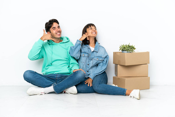 Young couple making a move while picking up a box full of things sitting on the floor isolated on white background making phone gesture. Call me back sign - Photo, Image