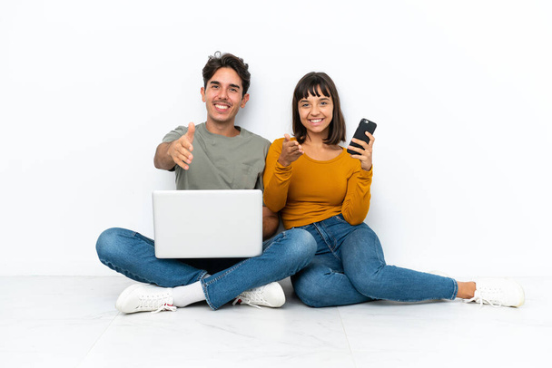 Jong stel met een laptop en mobiel zitten op de vloer handen schudden voor het sluiten van een goede deal - Foto, afbeelding