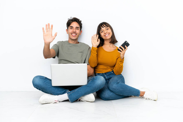 Young couple with a laptop and mobile sitting on the floor saluting with hand with happy expression - Photo, image
