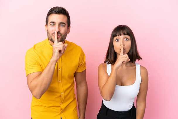 Young couple isolated on pink background showing a sign of silence gesture putting finger in mouth - Photo, Image