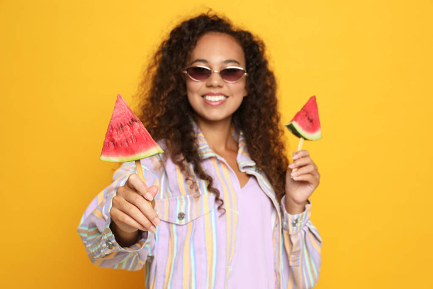 Beautiful young African American woman with pieces of watermelon against yellow background, focus on hand - Fotografie, Obrázek