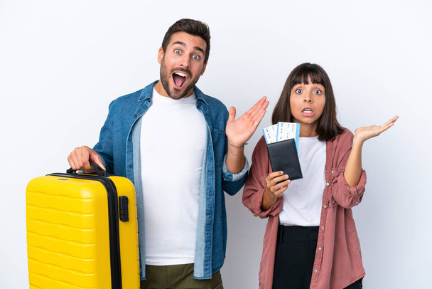 Joven pareja de viajeros sosteniendo una maleta y pasaporte aislados sobre fondo blanco con expresión facial sorpresa y conmocionada - Foto, imagen