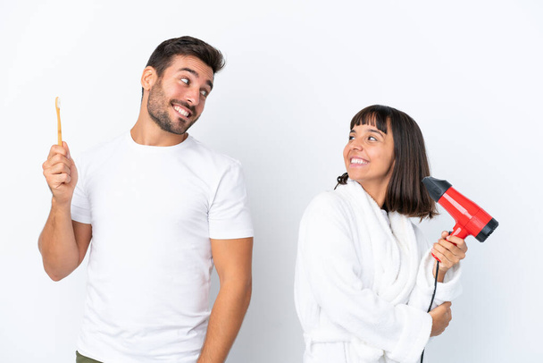 Jovem casal caucasiano segurando um secador de cabelo e escova de dentes isolado no fundo branco olhando sobre o ombro com um sorriso - Foto, Imagem