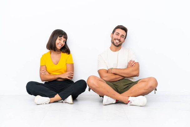 Jeune couple assis sur le sol isolé sur fond blanc gardant les bras croisés en position latérale tout en souriant - Photo, image