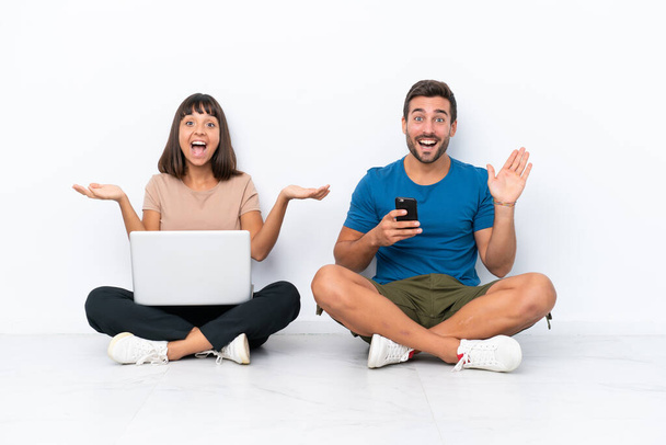 Молодая пара сидит на полу держа компьютер и мобильный телефон изолированы на белом фоне с удивлением и шокированным выражением лица - Фото, изображение