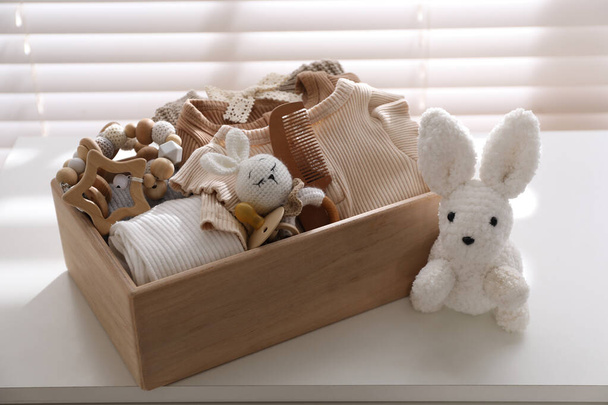 Wooden crate with children's clothes, toys and pacifier on white table near window - Fotó, kép