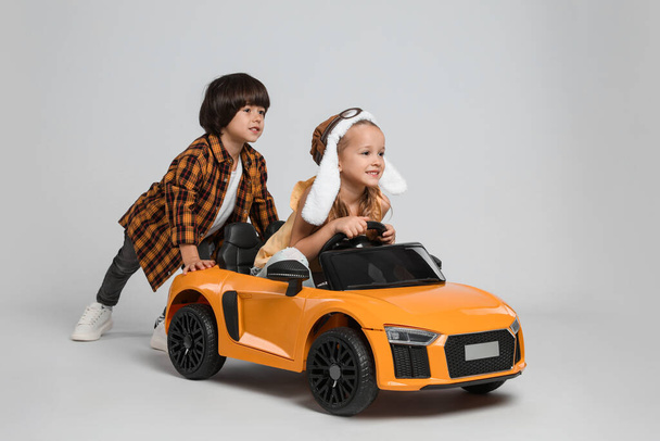 Cute boy pushing children's electric toy car with little girl on grey background - Fotoğraf, Görsel