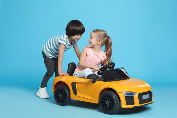 Cute boy pushing children's electric toy car with little girl on light blue background - 写真・画像