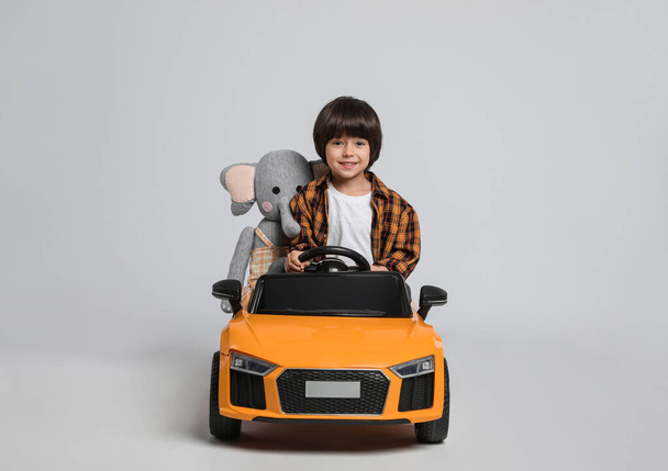 Cute little boy with toy elephant driving children's car on grey background - Φωτογραφία, εικόνα