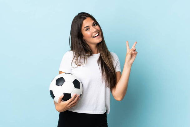 Young football player brazilian girl isolated on blue background smiling and showing victory sign - Foto, imagen