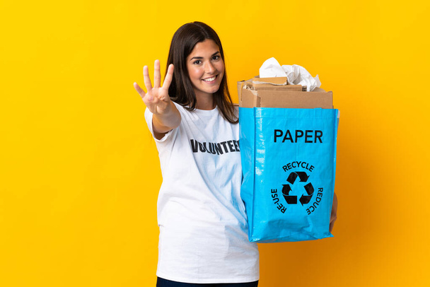 Νεαρή Βραζιλιάνα που κρατάει μια σακούλα ανακύκλωσης γεμάτη χαρτί για να ανακυκλώνει απομονωμένη σε κίτρινο φόντο χαρούμενη και μετρώντας τέσσερα με δάχτυλα. - Φωτογραφία, εικόνα