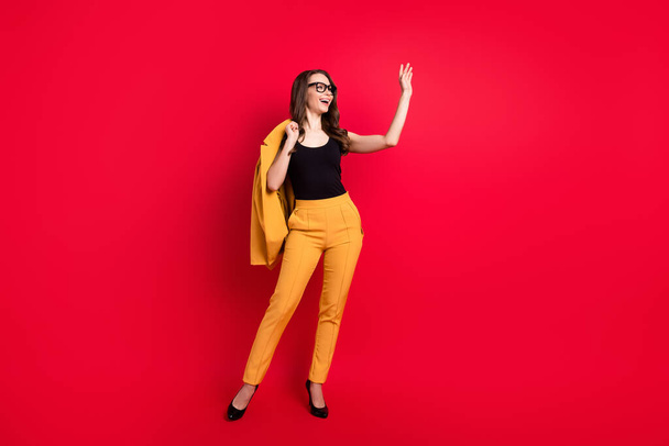 Photo of pretty charming young woman wear yellow jacket spectacles waving arm empty space smiling isolated red color background - 写真・画像
