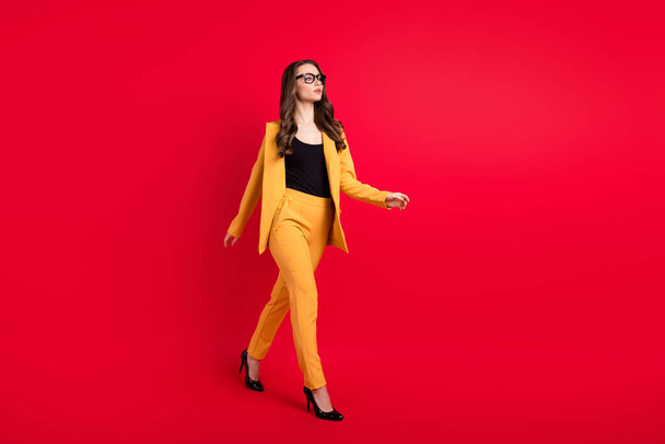 Photo of shiny serious young woman wear yellow jacket spectacles walking empty space isolated red color background - Foto, afbeelding