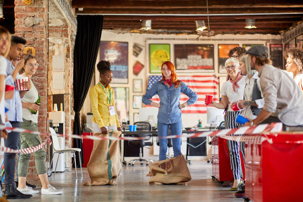 caucasian redhead and afroamerican female colleagues are about to race in sacks at work, with other employees cheering - 写真・画像