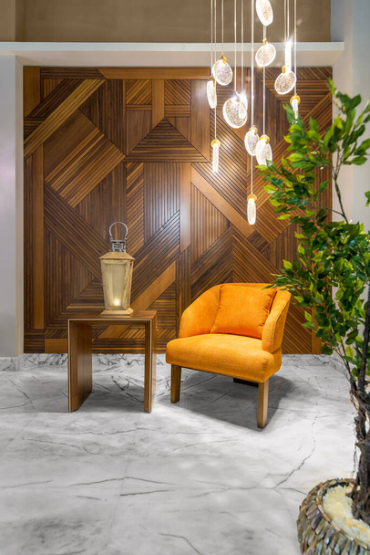 Sillón naranja moderno, linterna de madera en una mesa pequeña, candelabro de cristal alto contemporáneo y pared de revestimiento de madera. - Foto, Imagen