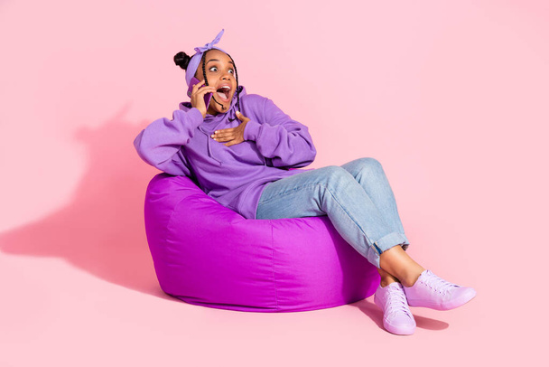 Fotografie atraktivní krásné africké dáma oblečené fialové mikina buchty mluví moderní gadget sedí fazole taška izolované růžové barvy pozadí - Fotografie, Obrázek