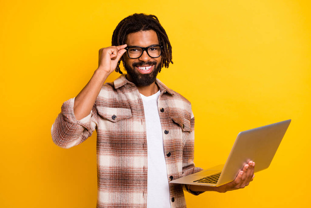 Photo portrait of brunet man touching glasses keeping laptop smiling doing project isolated on vibrant yellow color background - Фото, зображення