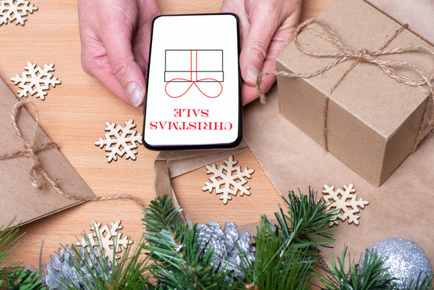 Close up hands holding a mobile phone with a white screen with the inscription SALE and a drawn gift on a wooden table with Christmas gift boxes, snowflakes, Christmas tree. Buying gifts for Christmas - Foto, immagini