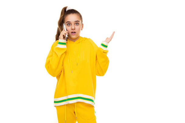 Photo of a beautiful shocked and surprised girl with dark hair with a ponytail in a stylish yellow tracksuit with green stripes talking on a mobile phone isolated on white background with free space - Photo, Image