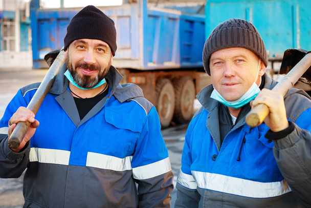 Двое рабочих в рабочей одежде и медицинских масках стоят с лопатами на улице и смотрят в камеру. - Фото, изображение