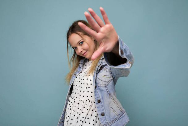 Photo of young positive attractive blonde woman with sincere emotions wearing jean blue jacket isolated over blue background with free space and showing no gesture with hand palm. Stop gesture - Zdjęcie, obraz