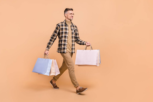 Full size profile side photo of mature man hold bag walk look empty space boutique isolated over beige color background - Fotoğraf, Görsel