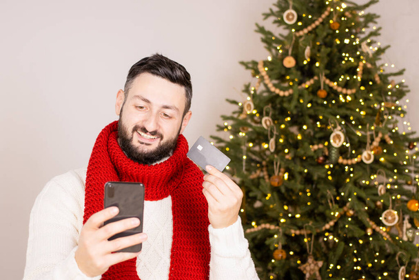 Щаслива брюнетка бородатий чоловік платить в інтернеті за допомогою банківської кредитної картки на різдвяні подарунки, тримаючи смартфон, дивлячись на екран, стоячи біля святкового дерева вдома, в червоному шарфі. Шопінг онлайн
. - Фото, зображення