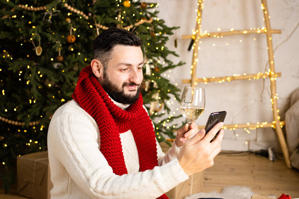 Smiling brunette bearded man making video call with smartphone, wearing red scarf, sitting near christmas tree indoor. Best friends toasting drinking champagne. Video conference party online meeting - Fotó, kép